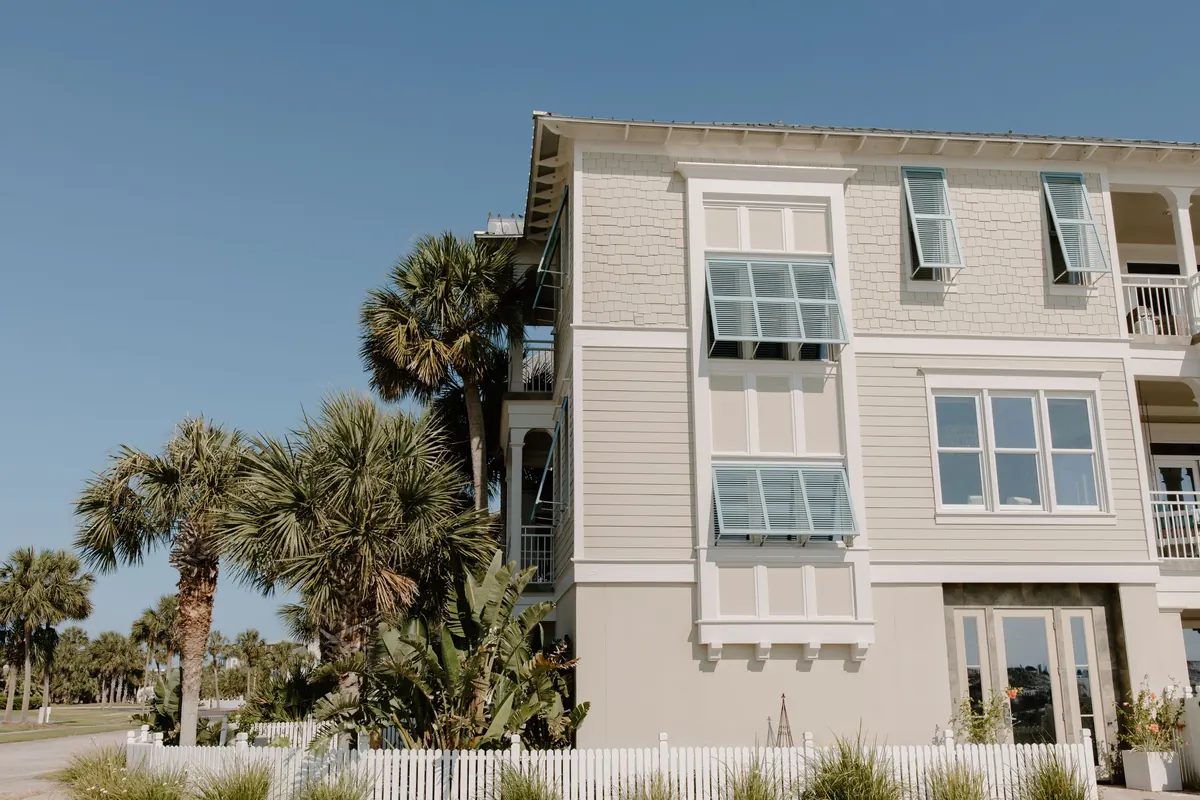 Light gray coastal home with white trim, Bahama shutters, palm trees and a white picket fence