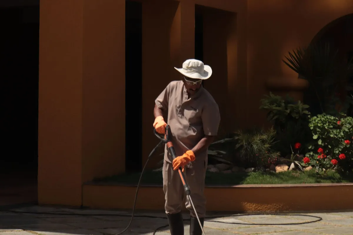 Worker in a sun hat and orange gloves pressure washing a paved walkway